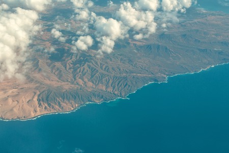 Idyllic azure coast with bald mountain peaks or brown tops under soft white clouds on blue sea water background. Aerial view from plane flightの写真素材