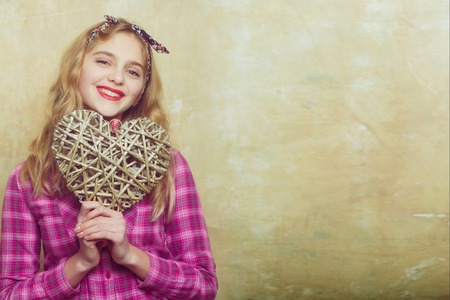 Happy pretty girl or beautiful woman with blond curly hair in stylish headband and violet plaid shirt smiles with wicker heart for valentines day on beige wall.の写真素材