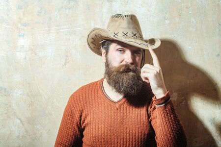 Bearded man, caucasian hipster, with long beard and moustache wearing cowboy hat and brown sweater poses with serious face on beige wall background, copy spaceの写真素材