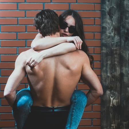 young couple of handsome macho man with muscular back and hands holds pretty woman or sexy girl with long hair in fashionable sunglasses and jeans on brick wall backgroundの写真素材