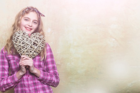 Happy pretty girl or beautiful woman with blond curly hair in stylish headband and violet plaid shirt smiles with wicker heart for valentines day on beige wall background, copy spaceの写真素材