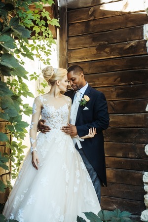 Happy smiling couple of handsome man or african American groom hugging pretty girl or beautiful bride in white wedding dress among green vine leaves on sunny summer day on wooden wall backgroundの写真素材