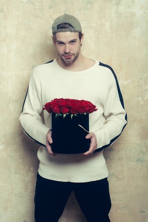 bearded macho smiling man, brutal caucasian hipster with moustache in sweater and cap holds red rose...