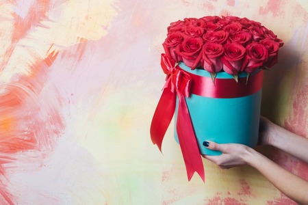 Beautiful red roses, bunch of fresh natural flowers, in blue present box with ribbon in female hands of woman on colorful abstract background, copy spaceの写真素材
