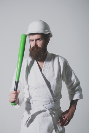 bearded karate man, long beard, brutal caucasian hipster with moustache in white kimono with black belt, builder helmet holds green baseball bat on grey background, unshaven guyの写真素材