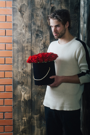 bearded macho man, brutal caucasian hipster with moustache in sweater holds red rose flowers in box as valentines day present on wooden and brick wall backgroundの写真素材