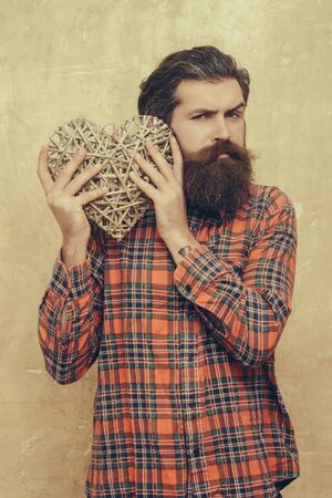 bearded man, caucasian hipster, with long beard and moustache in plaid shirt holds wicker heart, love gift for valentines day, shouting on beige grunge wall backgroundの写真素材