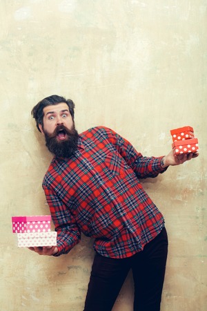 surprised bearded man, caucasian hipster, with long beard and moustache in red plaid shirt holding colorful gift boxes stacked in hands on beige wall backgroundの写真素材