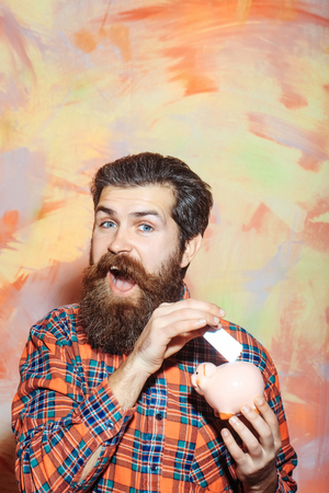 Happy bearded man, caucasian hipster, with long beard and moustache in red plaid shirt putting banknote in pink ceramic piggy bank on abstract colorful wall background. Saving money conceptの写真素材