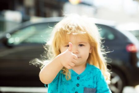 Cute baby boy with long blond hair gives thumbs up hand gesture walking on city street on sunny summer day outdoors on blurred backgroundの写真素材
