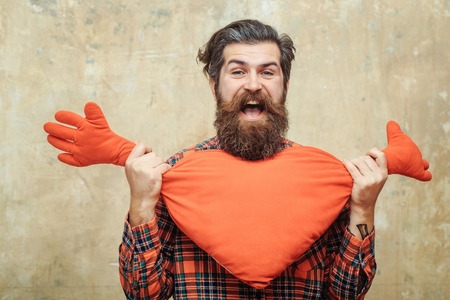 Happy bearded man, caucasian hipster, with long beard and moustache in plaid shirt holding red heart shape toy with hands on beige wall background. Love gift for valentines dayの写真素材