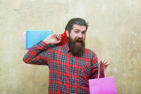 Happy bearded man, caucasian hipster, with long beard and moustache in red plaid shirt holding colorful paper shopping bags on beige wall backgroundの写真素材