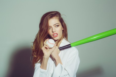 young pretty woman or cute sexy girl with beautiful long hair and red lips on adorable face in fashionable white shirt holds green baseball bat or racket and ball on grey backgroundの写真素材