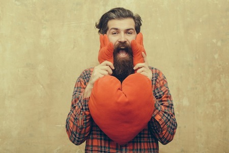 Happy bearded man, caucasian hipster, with long beard and moustache in plaid shirt holding red heart...