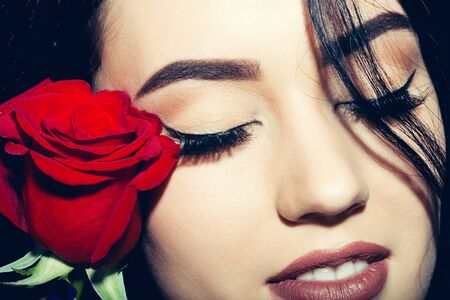 Pretty smiling girl or beautiful woman with brunette hair and stylish makeup on young face with closed eyes and red rose, natural fresh flower, on blue backgroundの写真素材