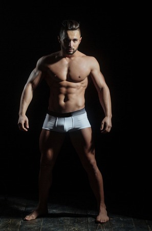 handsome muscular macho man or athlete guy with sexy body of bodybuilder with torso, belly, six packs, has beard on serious face in white pants isolated on black background on wooden floorの写真素材