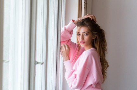 Pretty girl or beautiful woman with long, blond hair, hairstyle, in cute, rosy sweater standing at window on white wall backgroundの写真素材