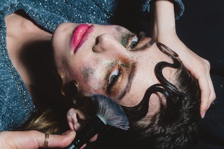 Pretty girl or fashion woman putting holiday, glitter makeup, sparkling eyeshadow, rosy lips, with make up brush and cute, curly lock hair, hairstyle, on young face on dark backgroundの写真素材