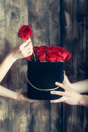 Beautiful red roses, bunch of fresh natural flowers, in black present box in human hands of man and woman on wooden wall backgroundの写真素材