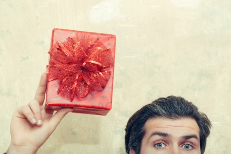 Beautiful red gift box with bow, present holidays package, in hand of caucasian man with blue eyes and brunette hair with grey on beige wall backgroundの写真素材