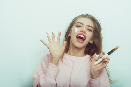 happy pretty girl or beautiful woman with long, blond hair, putting facial cream or mask on smiling face skin with brush from jar on white background. Cosmetology, beauty and spa, copy spaceの写真素材