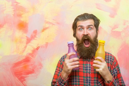 surprised bearded man, caucasian hipster, with long beard and moustache in red plaid shirt holding two plastic bottles, yellow and violet color, on abstract pink wall backgroundの写真素材
