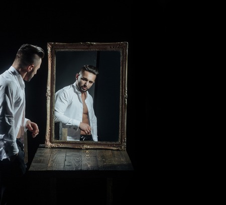 handsome muscular macho man or athlete guy with sexy body of bodybuilder with torso, belly, six packs, has beard on serious face in pants, white shirt near vintage mirror isolated on black backgroundの写真素材