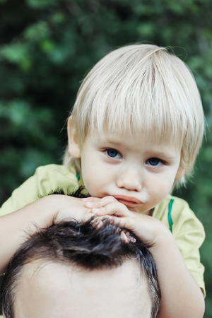 Tired cute baby boy, son, little child, with blond hair sitting on father neck or shoulders on sunny, summer day outdoors on natural backgroundの写真素材