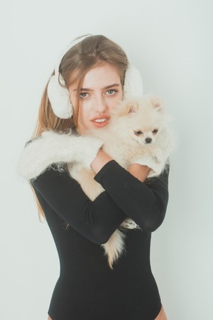 pretty girl or sexy woman with long blond hair, hairstyle and adorable face in black bodysuit, gloves, mittens, earmuffs holds small cute pomeranian spitz dog or puppy pet isolated on white backgroundの写真素材