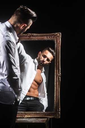 handsome muscular macho man or athlete guy with sexy body of bodybuilder with torso, belly, six packs, has beard on serious face in pants, white shirt near vintage mirror isolated on black backgroundの写真素材