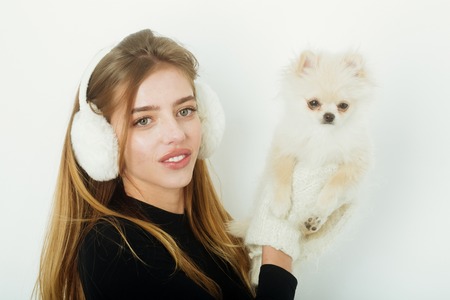 pretty girl or sexy woman with long blond hair, hairstyle and happy face in black bodysuit, gloves, mittens, earmuffs holds small cute pomeranian spitz dog or puppy pet isolated on white backgroundの写真素材