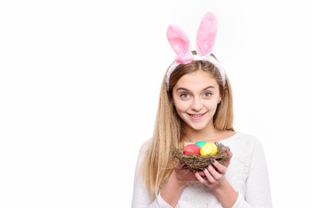 colorful painted easter eggs in straw nest and happy girl in pink bunny ears with long blonde hair and smiling adorable face isolated on white background. traditional spring holiday food, copy spaceの写真素材