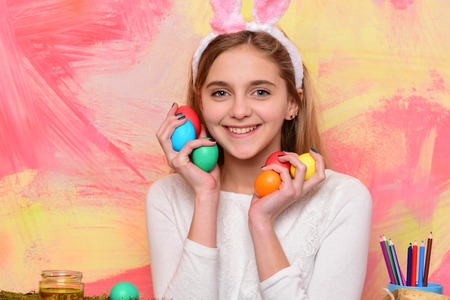 happy easter girl painter in pink bunny ears with colorful painted eggs and pencil set, has long blonde hair on abstract background. traditional spring holiday celebrationの写真素材
