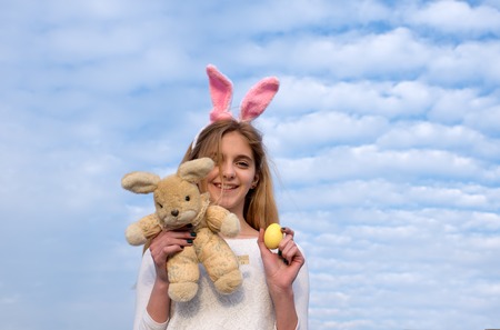 yellow painted easter egg as traditional spring holiday food and rabbit toy, happy girl in pink bunny ears with, has long blonde hair and smiling adorable face outdoor on blue cloudy sky backgroundの写真素材