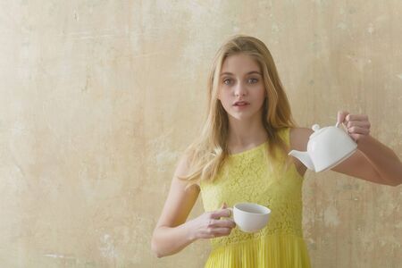 Pretty girl or cute woman with long blond hair and adorable face in yellow dress holds tea, coffee cup and teapot on textured wall background, copy spaceの写真素材