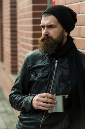 bearded man, long beard, brutal caucasian hipster with moustache holding metallic flask, jar, has serious face, in black leather jacket, hat, scarf on brick wall background outdoorの写真素材