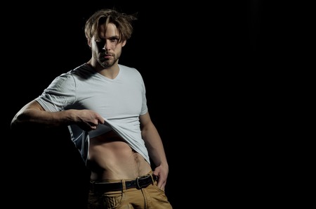 Handsome man or unshaven macho, bodybuilder, with stylish blond hair, haircut, showing sexy, muscular torso with six packs and abs, biceps, triceps in white tshirt, striptease on black backgroundの写真素材