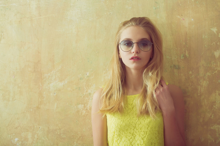 Pretty girl or cute woman with long blond hair and adorable face in yellow dress and sunglasses on textured wall background, copy spaceの写真素材