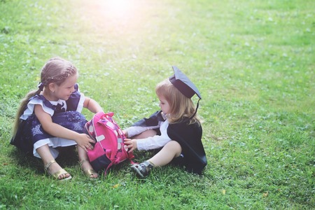 Cute little children, blond baby boy in black graduation hat or cap and robe and small girl with long hair with pink school bag, backpack, sitting on green grass on summer day on natural backgroundの写真素材