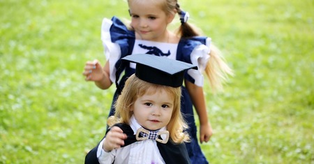 Cute little girl with long hair in blue dress dressing adorable small boy in black graduation hat or cap and robe on summer day outdoors on green grass backgroundの写真素材