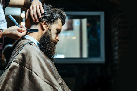 Bearded man with long beard, brutal, caucasian hipster with moustache, getting stylish hair shaving, haircut, with razor by barber or hairdresser hands at barbershopの写真素材