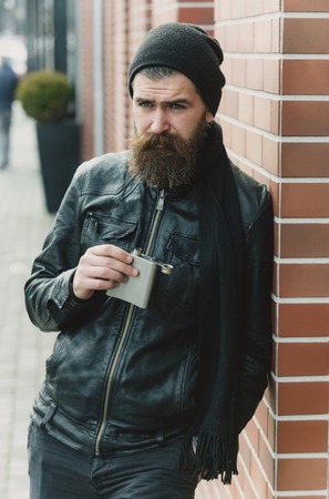 bearded man, long beard, brutal caucasian hipster with moustache holding metallic flask, jar, has serious face, in black leather jacket, hat, scarf on brick wall background outdoorの写真素材