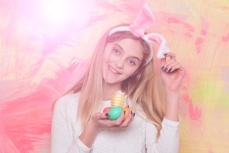 happy girl in pink bunny ears with long blonde hair and adorable smiling face holds colorful painted easter eggs on abstract background. traditional spring holiday celebrationの写真素材
