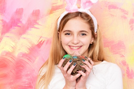 happy easter girl in pink bunny ears with colorful painted eggs in straw nest, has long blonde hair and smiling adorable face on abstract background. traditional spring holiday food, copy spaceの写真素材