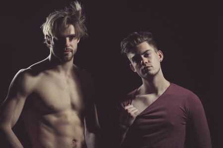 Two handsome men posing on black background. Muscular macho, unshaven caucasian, with muscle torso with six packs and abs, biceps, triceps. Male model in dark red tshirt with long sleeves, casual wearの写真素材