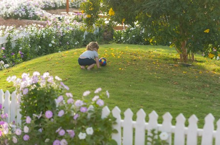 sunny summer day on natural background with cute baby boy, small, little child with long blond hair, ponytail, barefoot, playing with colorful ball on green grass in garden with blossoming flowersの写真素材