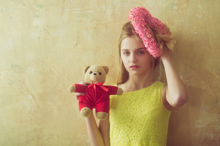 Pretty girl or cute woman with blond hair and adorable face in yellow dress with teddy bear toy and pink wicker heart for valentines day on textured wall background, copy spaceの写真素材