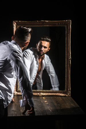 handsome muscular macho man or athlete guy with sexy body of bodybuilder with torso, belly, six packs, has beard on serious face in pants, white shirt near vintage mirror isolated on black backgroundの写真素材