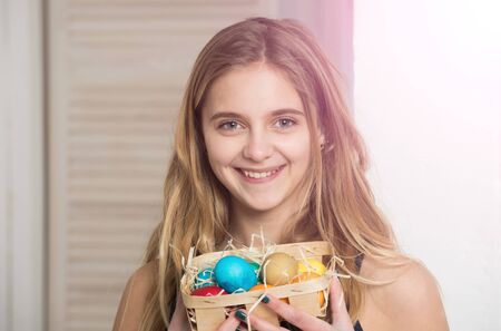 happy easter girl with colorful painted eggs in basket, has long blonde hair and smiling cute face on blurred background. traditional spring holiday food. celebration and decorationの写真素材