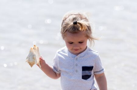 happy cute baby boy, small, little child with blond hair smiling with seashell or marine shell in hand on sunny day on bokeh sea water background. Idyllic summer vacationの写真素材
